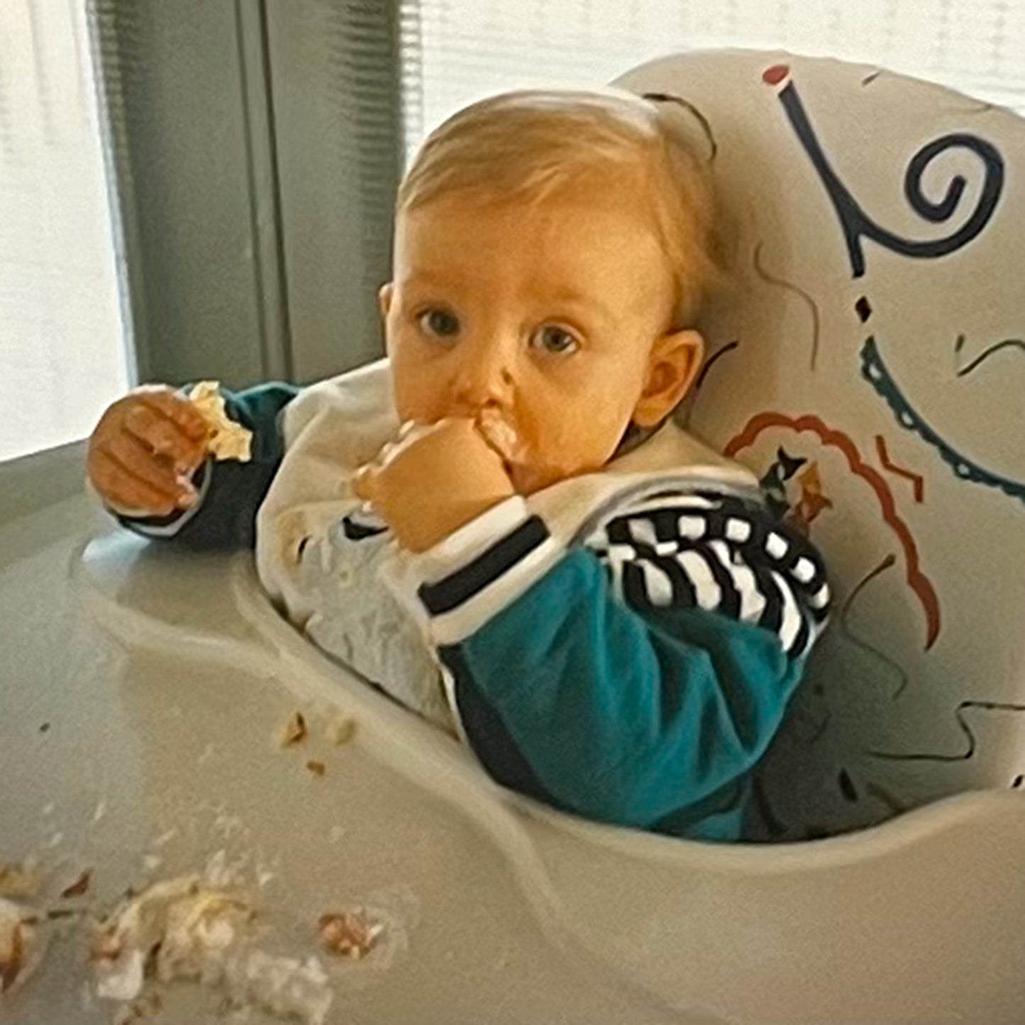 Blonde baby in highchair is eating cake with his hands