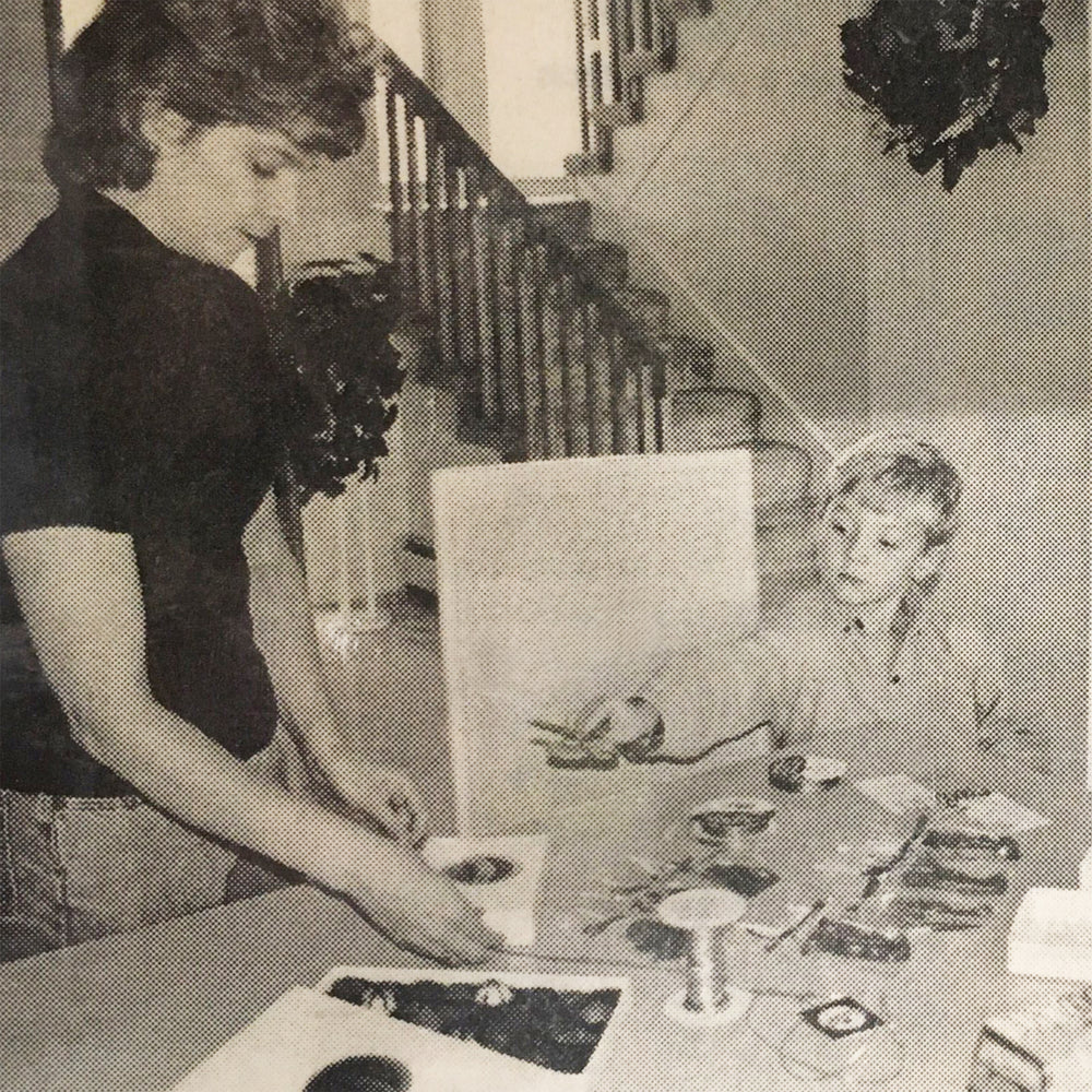 Woman and young boy working at a table. Woman has a box of truffles. Child is handing woman a pair of scissors.