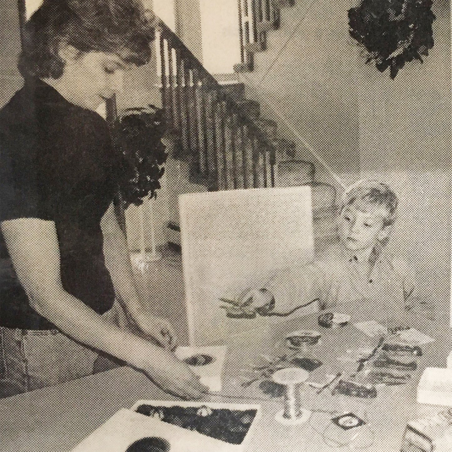 Black and white picture of mother and son packing boxes of chocolate. Son hands mother scissors to cut ribbon.