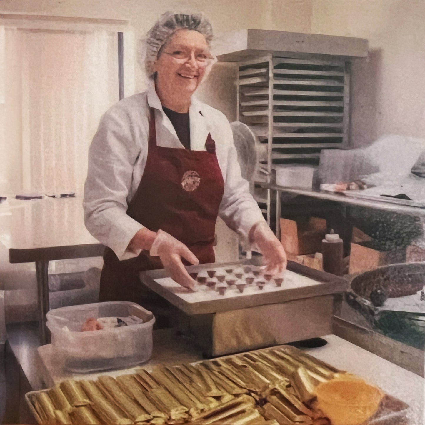 Old woman in glasses smiling at camera holding a tray of chocolate drops in production kitchen