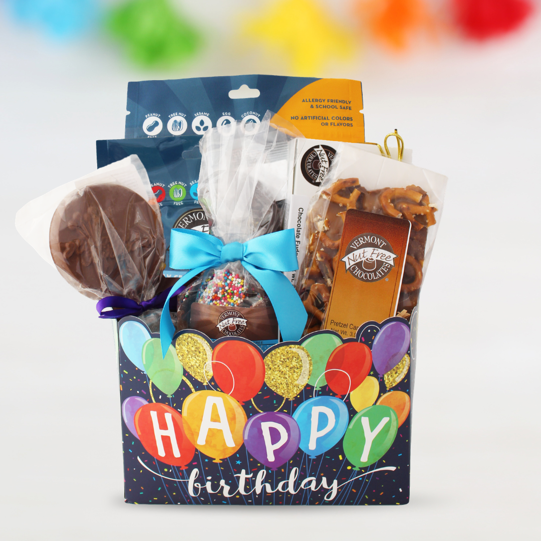 A chocolate and snack gift basket in a dark blue 'HAPPY birthday' balloon-themed box, filled with assorted packaged sweets, pretzels, and a chocolate-covered ice cream cone, set against a blurred background with colorful decor.