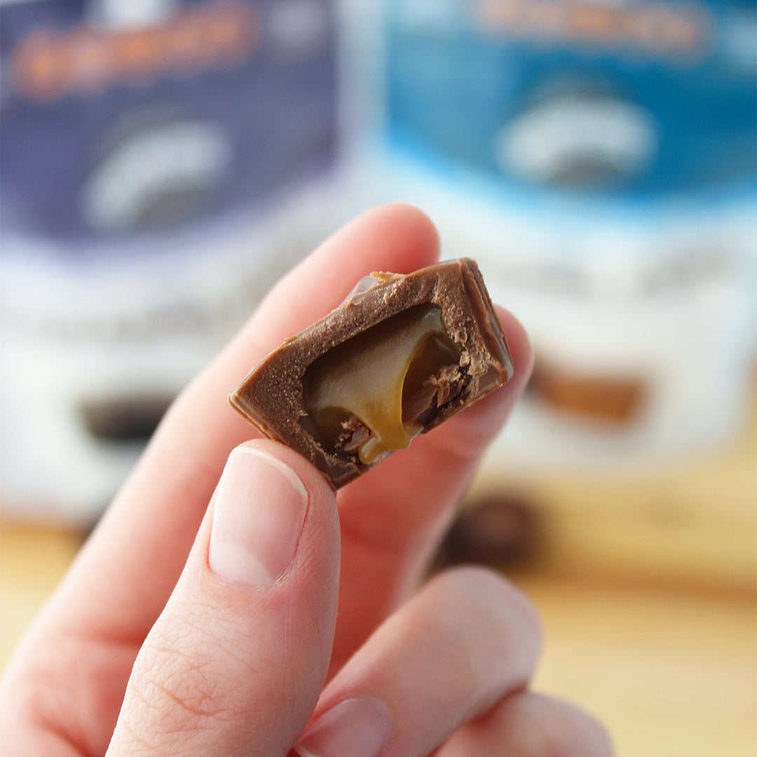 A close-up of a hand holding a broken chocolate candy with a caramel filling, with blurred chocolate packages in the background.