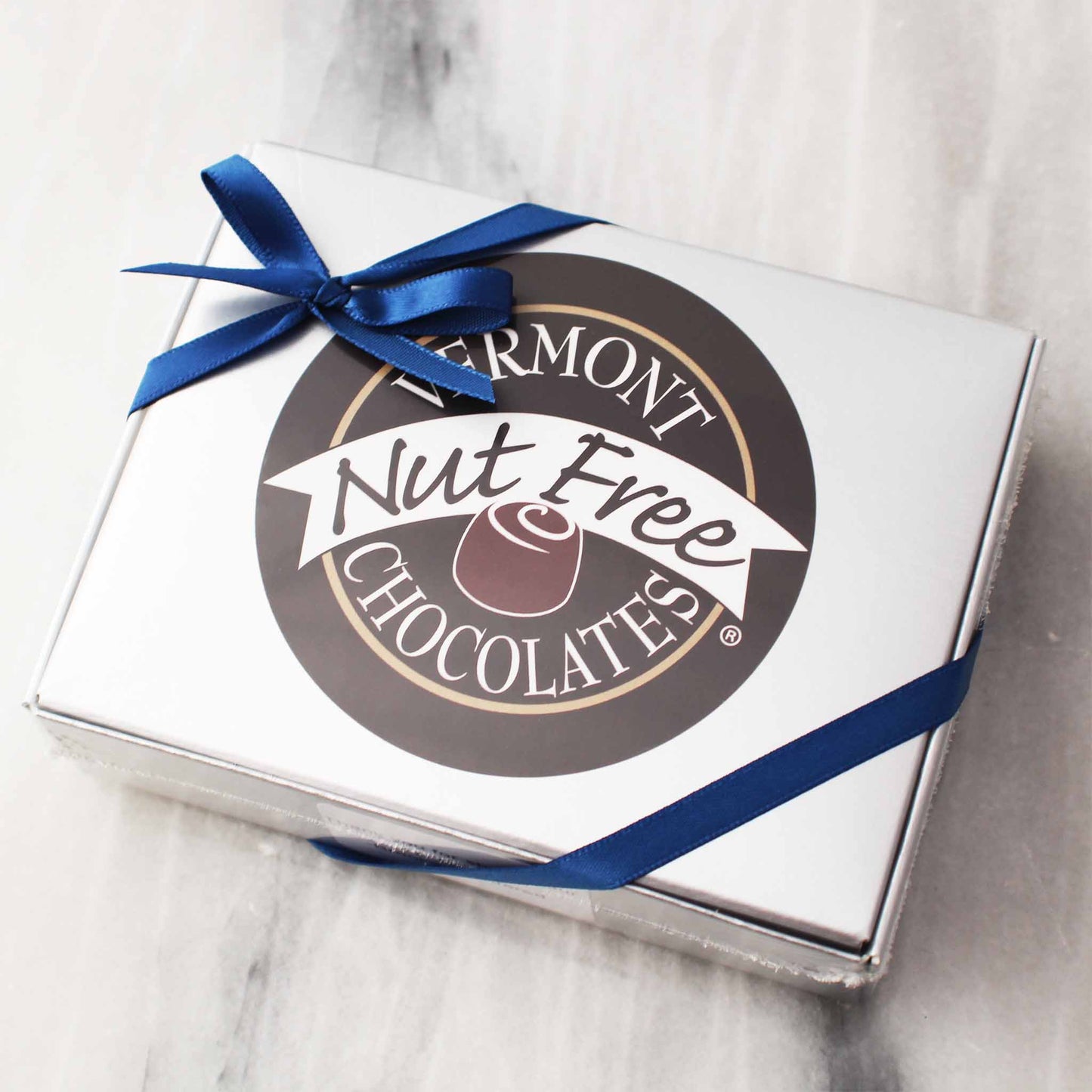 Silver box with a blue ribbon. Big circular logo on the front of the box that reads, "Vermont Nut Free Chocolates"