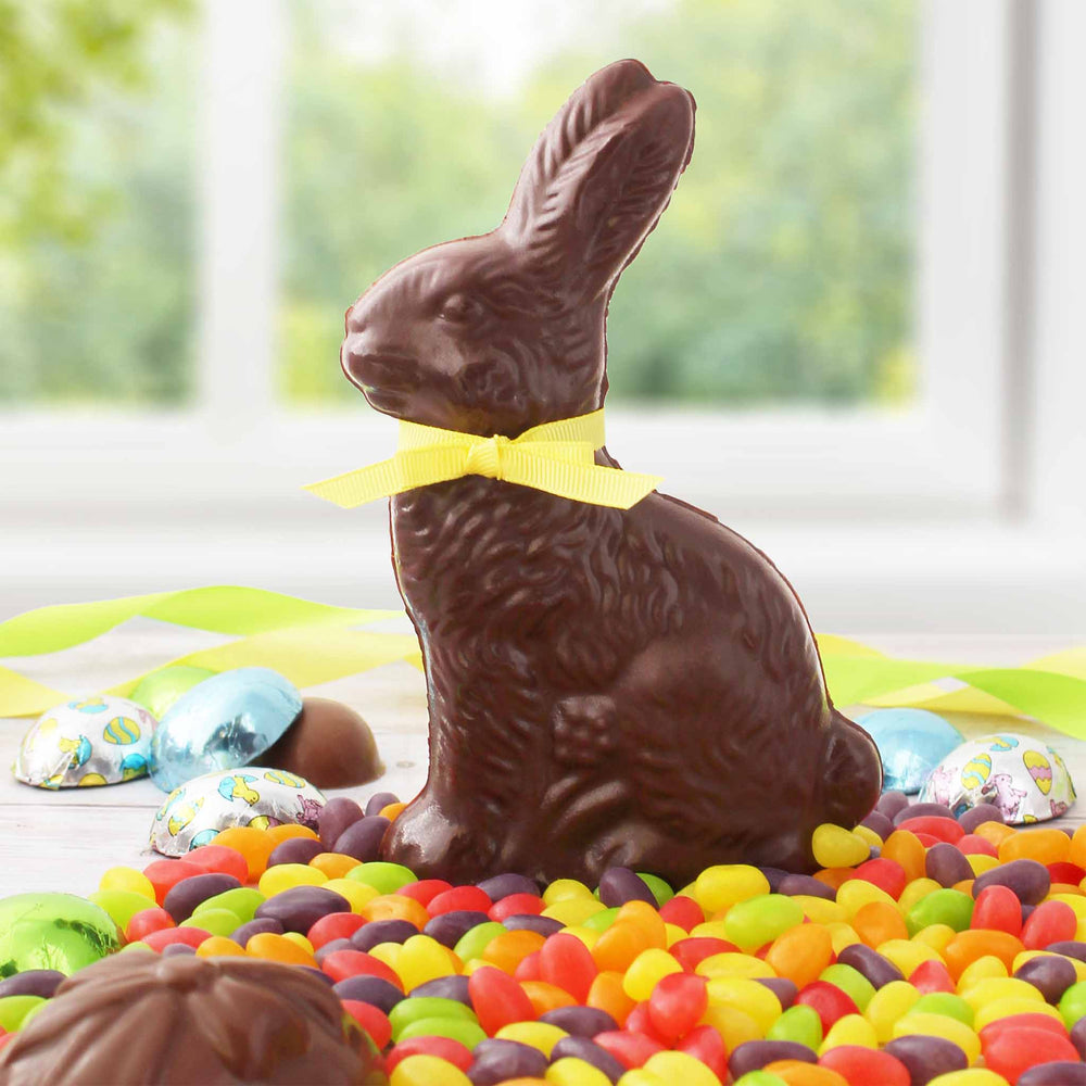 Chocolate Easter bunny with yellow ribbon tied around its neck. Sitting on a table filled with jelly beans. Chocolate eggs visible in background.