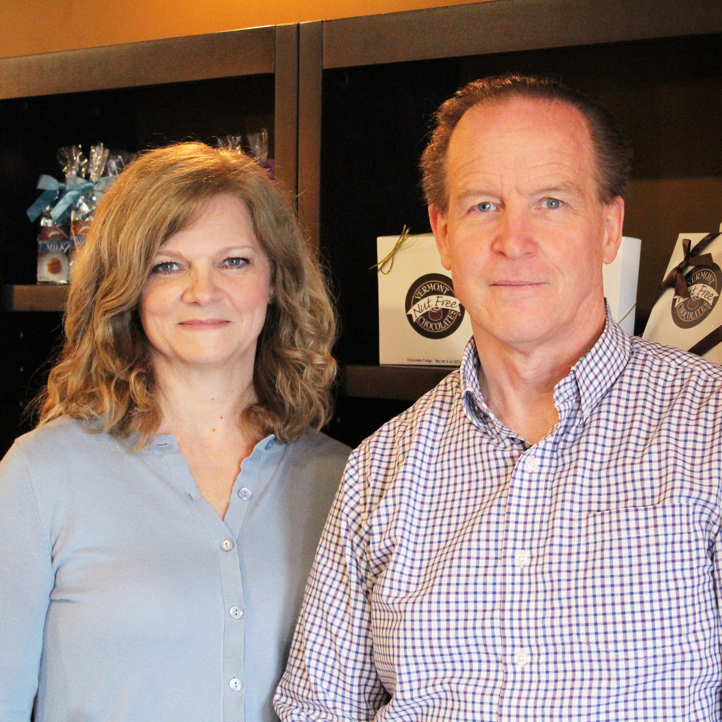 Headshot of man and woman standing side by side, smiling. Vermont Nut Free Chocolates boxes in the background on shelf.