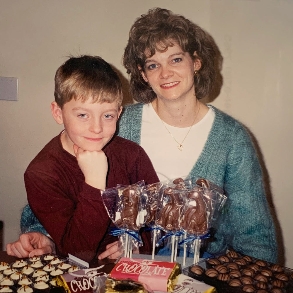 Woman and small boy sitting behind a table of chocolates. Boy is sitting on womans lap, his head resting on his hand.