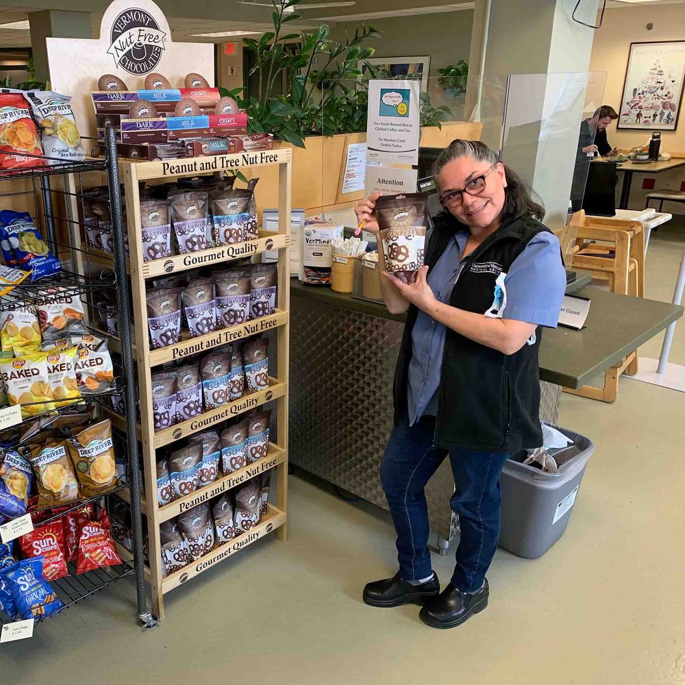 Woman standing in front of Vermont Nut Free Chocolates branded display rack. She is holding a bag of chocolate covered pretzels. The rack is filled with similar bags.