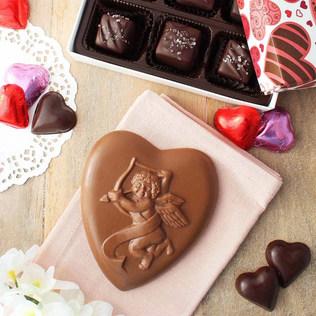 Assorted chocolates on wooden table top. Surrounded by flowers, doilies, and hearts. 