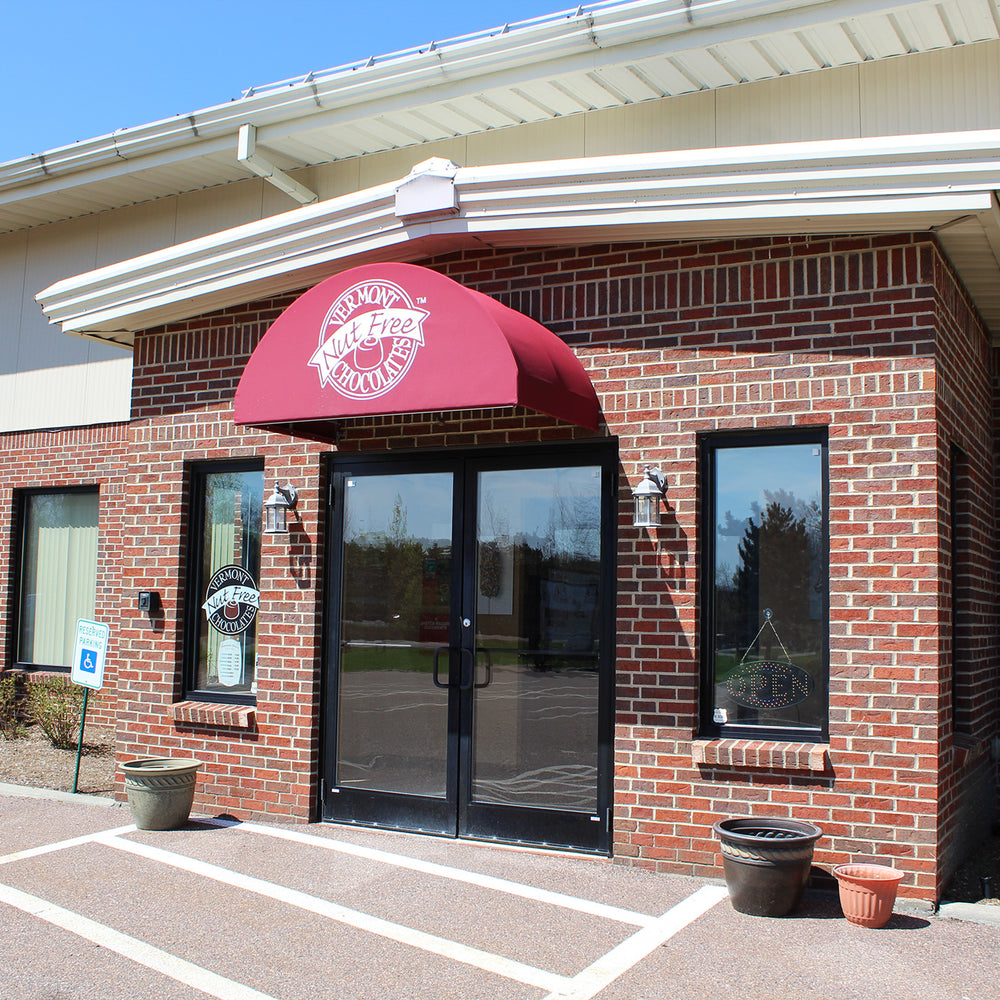 Front of Vermont Nut Free Chocolates factory store. Brick building with red awning. White Vermont Nut Free Chocolates logo on awning.